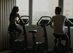 Salle de fitness vue en contre-jour avec une femme à gauche et un homme à droite pédalant sur des vélos d'appartement face à une grande fenêtre, un mur à lattes en bois clair à gauche, et une selle de vélo vide visible au premier plan au centre, idéal pour un séminaire bien-être.