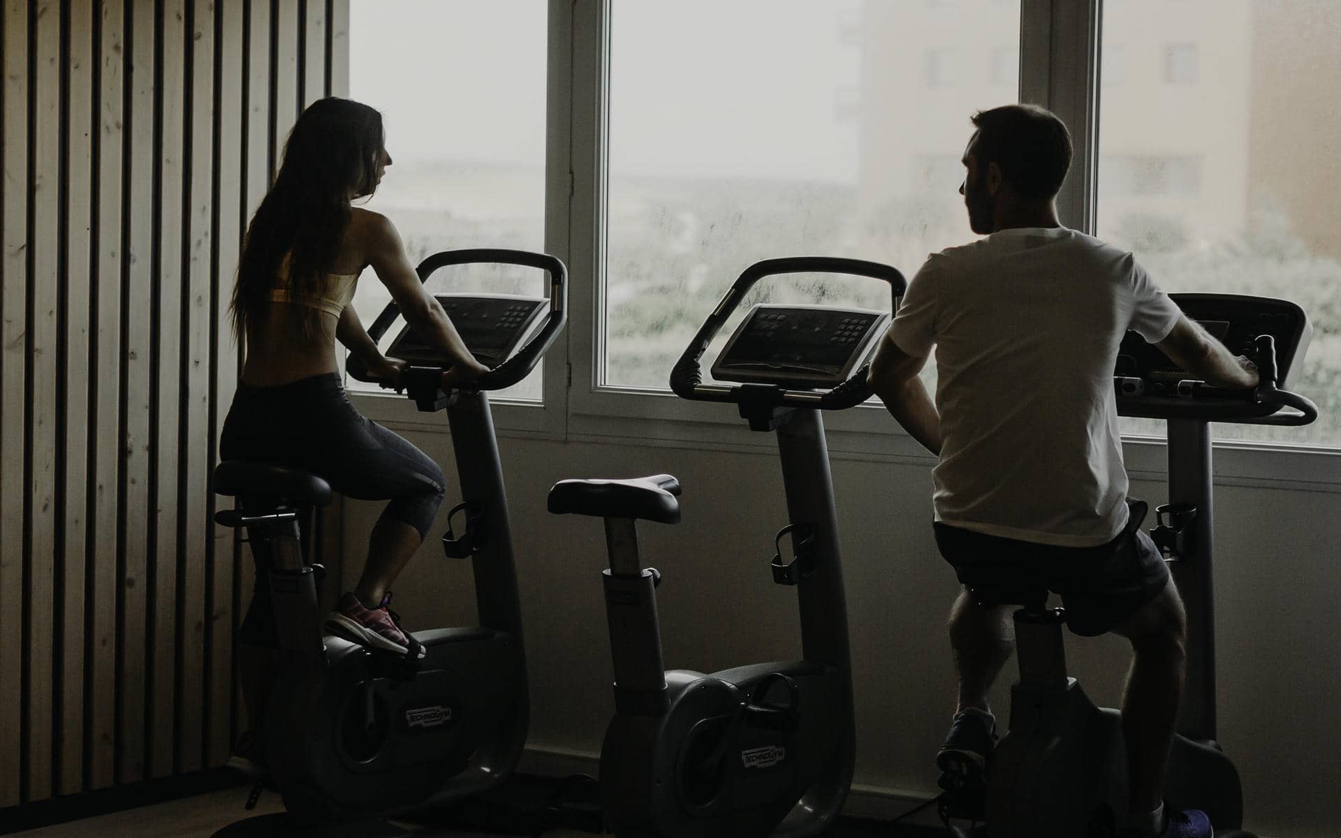 Salle de fitness vue en contre-jour avec une femme à gauche et un homme à droite pédalant sur des vélos d'appartement face à une grande fenêtre, un mur à lattes en bois clair à gauche, et une selle de vélo vide visible au premier plan au centre, idéal pour un séminaire bien-être.