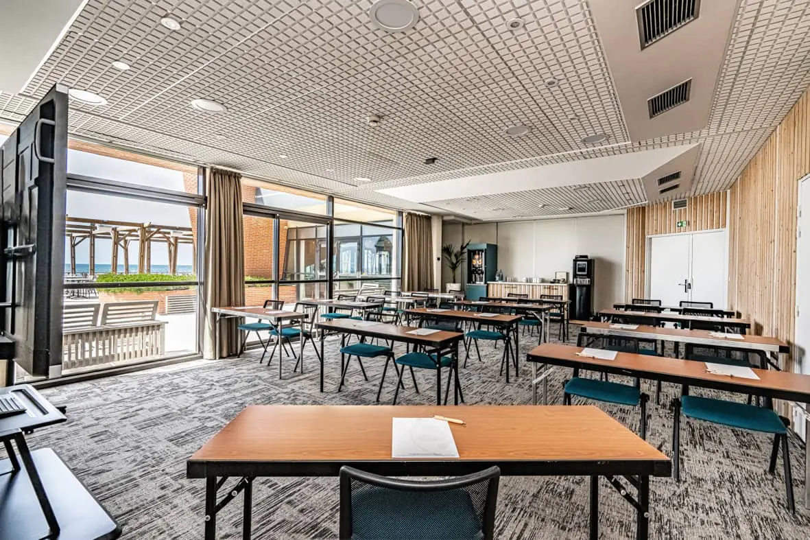 Salle de réunion aménagée en rangées façon théâtre avec des tables pliantes en bois et des chaises en tissu bleu canard à structure métallique noire, un écran de télévision noir au premier plan à gauche, des distributeurs de café et d'eau le long d'un mur à droite près d'un lambris en bois clair à lattes, un sol en moquette grise à motifs, et des baies vitrées du sol au plafond avec rideaux beiges laissant voir une vue sur la mer avec une pergola en bois à l'extérieur, idéal pour un séminaire en bord de mer.