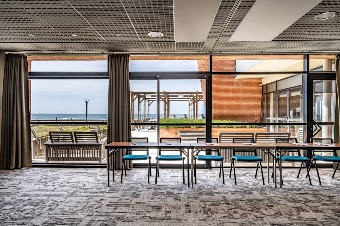 Salle de réunion avec une longue table pliante en bois et des chaises en tissu bleu canard à structure métallique noire face à l'objectif au centre, un sol en moquette grise à motifs, des rideaux marron encadrant les baies vitrées de part et d'autre, et une vue panoramique sur la mer avec une pergola en bois et une terrasse visibles à l'extérieur, idéal pour un séminaire en bord de mer.