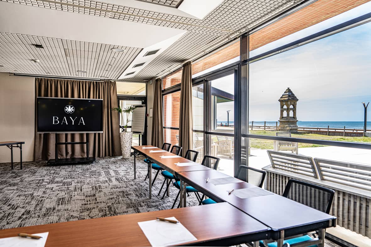 Salle de réunion avec des tables pliantes en bois disposées en ligne accompagnées de chaises en tissu bleu canard à structure métallique noire au premier plan, un écran numérique noir affichant un logo sur un support mobile avec des rideaux marron à gauche, un sol en moquette grise à motifs, et de grandes baies vitrées à droite offrant une vue panoramique sur la mer avec un phare en pierre et une terrasse de plage à l'extérieur, idéal pour un séminaire en bord de mer.