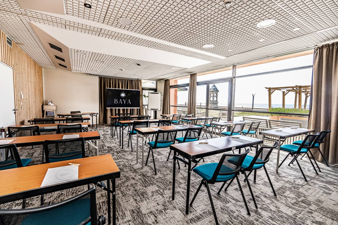 Salle de réunion aménagée en rangées façon théâtre avec des tables pliantes en bois et des chaises en tissu bleu canard à structure métallique noire, un écran numérique noir affichant un logo sur le mur en arrière-plan, un sol en moquette grise à motifs, et des baies vitrées du sol au plafond avec rideaux beiges offrant une vue panoramique sur la mer avec un phare en pierre à l'extérieur, idéal pour un séminaire en bord de mer.