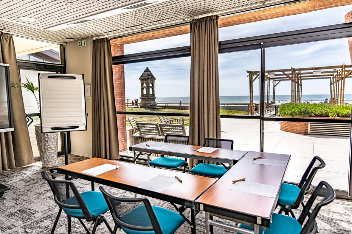 Salle de réunion avec des tables pliantes en bois disposées en U et des chaises en tissu bleu canard à structure métallique noire au premier plan, un paperboard blanc sur pied et un écran à gauche, un sol en moquette grise à motifs, des rideaux beiges encadrant les baies vitrées du sol au plafond, et une vue panoramique sur la mer avec un phare en pierre et une pergola en bois visibles à l'extérieur, idéal pour un séminaire en bord de mer.