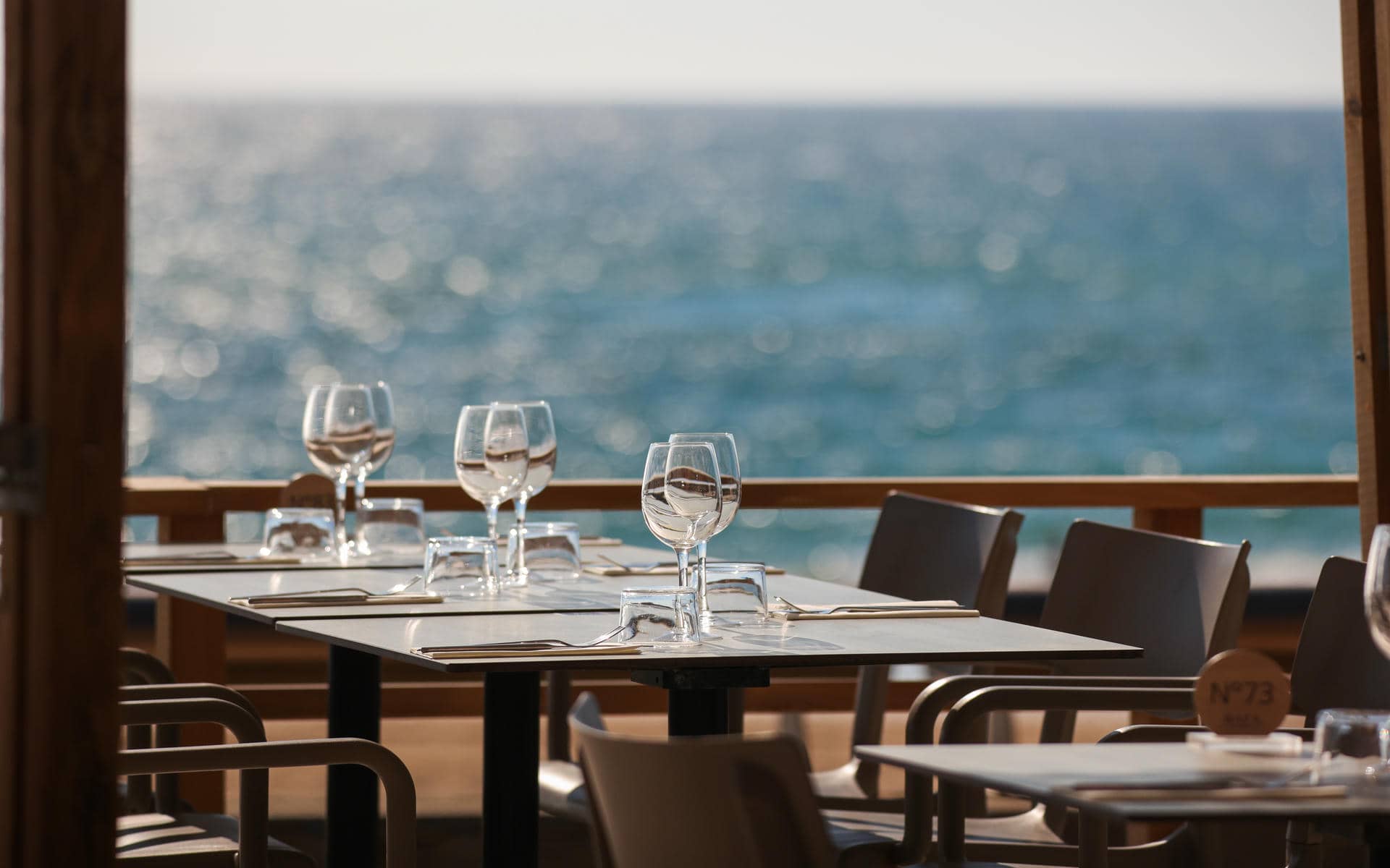 Restauration sur place gros plan sur une table grise dressée de verres à vin et à eau, serviettes et couverts au premier plan, des chaises en bois et d'autres tables dressées à droite, et une vue sur la mer floue visible au-delà d'une balustrade en bois en arrière-plan.