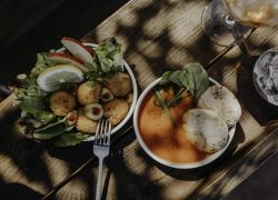 Vue de dessus d'un bol blanc avec des bouchées panées, des olives, des tranches de citron, de la pomme et de la salade à gauche, un bol blanc avec une soupe orange, des herbes et de fines galettes au centre, et des verres d'eau et de vin sur une table en bois en arrière-plan.