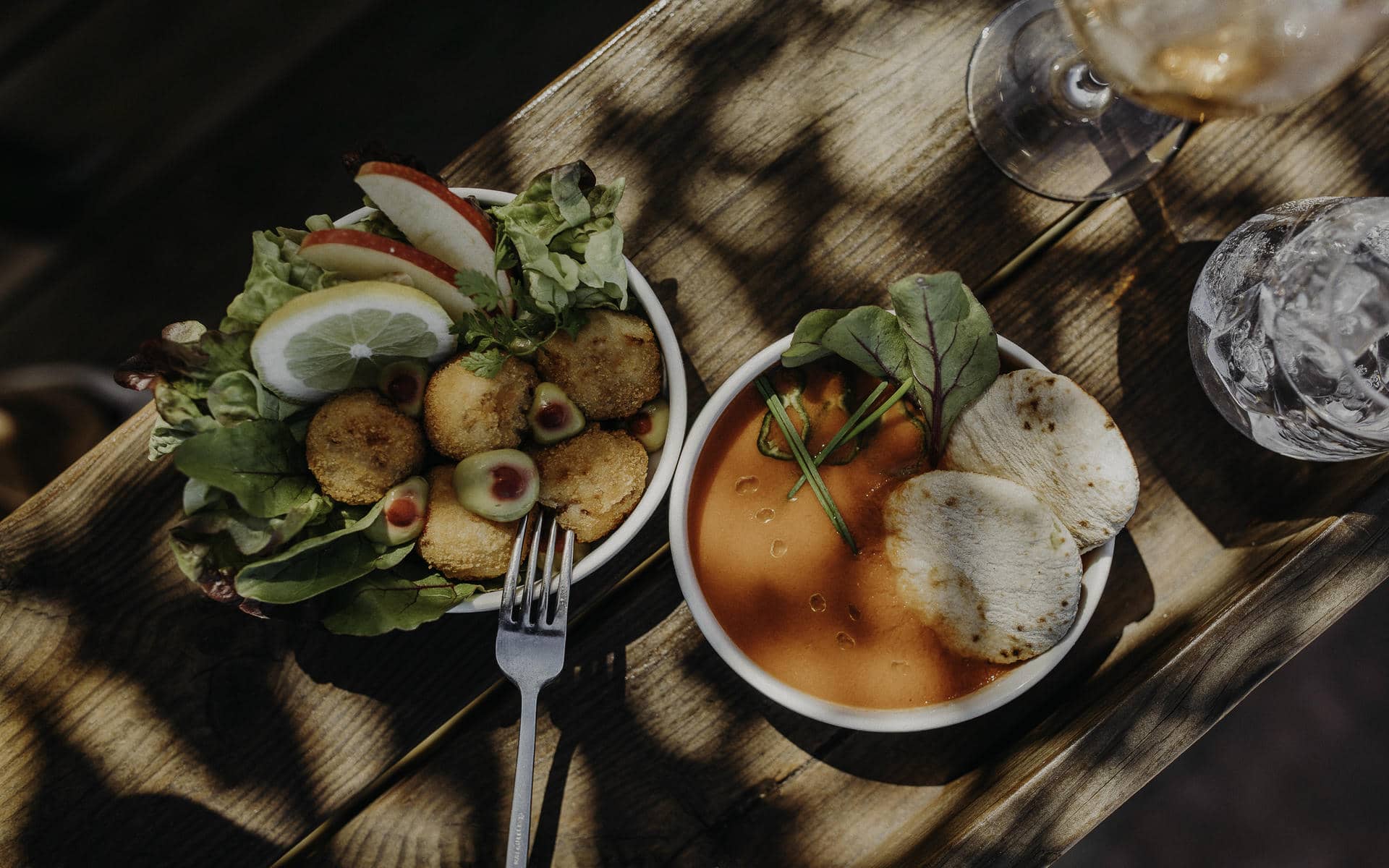 Vue de dessus d'un bol blanc avec des bouchées panées, des olives, des tranches de citron, de la pomme et de la salade à gauche, un bol blanc avec une soupe orange, des herbes et de fines galettes au centre, et des verres d'eau et de vin sur une table en bois en arrière-plan.