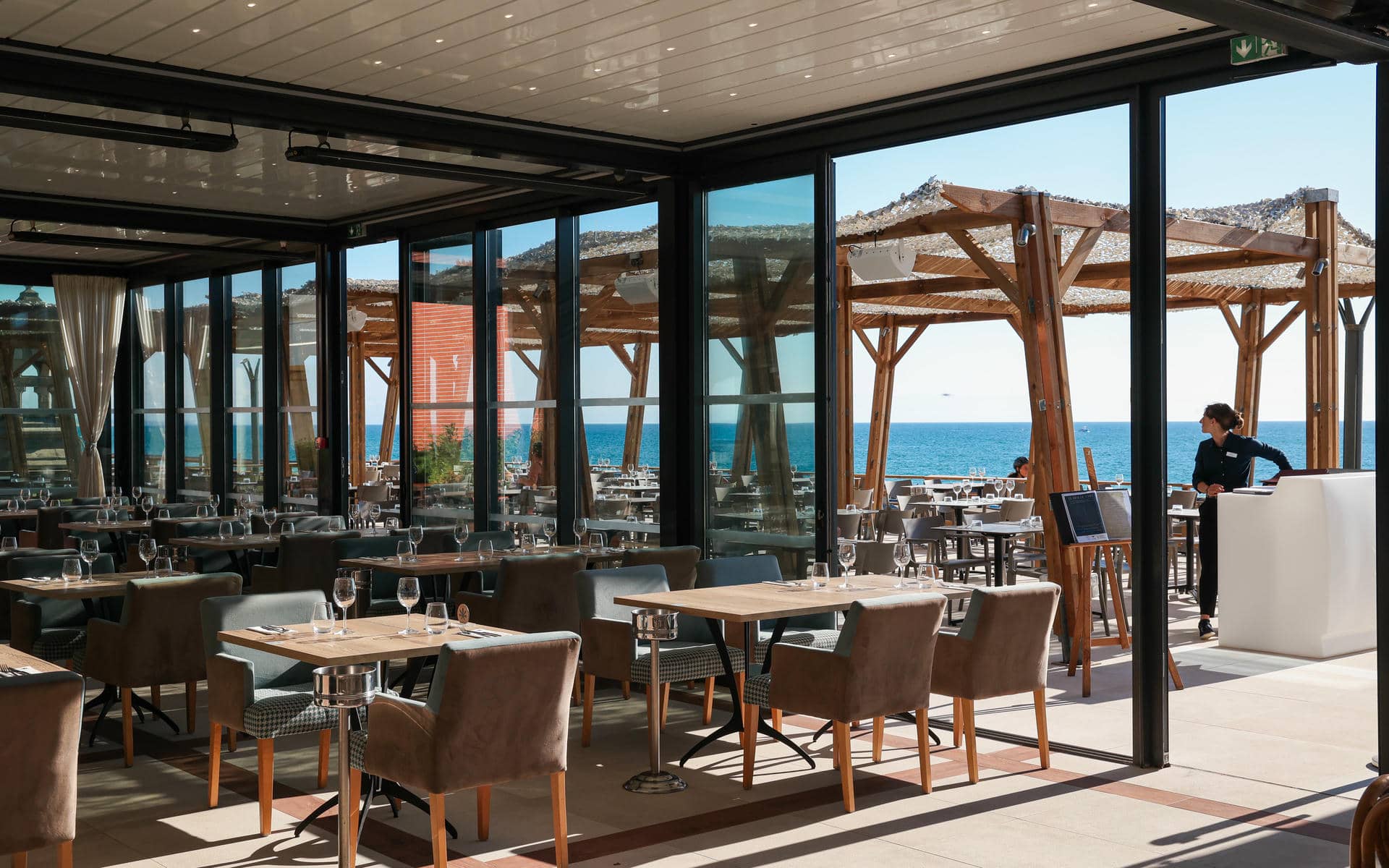 Restaurant sur place d'hôtel avec des tables en bois dressées de verres à vin et des chaises rembourrées grises et beige au premier plan, d'autres tables dressées et des seaux à glace sur pied métalliques au centre, et des baies vitrées du sol au plafond ouvrant sur une terrasse à pergola en bois avec vue mer et une personne debout près d'un comptoir d'accueil blanc en arrière-plan.