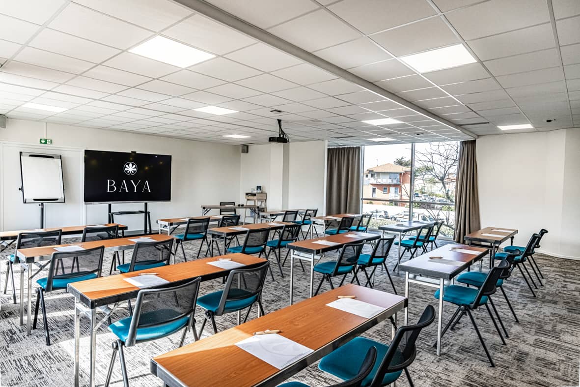 Salle de réunion aménagée en rangées façon théâtre avec des tables pliantes en bois et des chaises en tissu bleu canard à structure métallique noire, un grand paperboard à gauche et un écran numérique noir affichant un logo sur le mur en arrière-plan, un vidéoprojecteur fixé au plafond, un sol en moquette grise à motifs, et une grande fenêtre avec rideaux beiges laissant voir un quartier résidentiel à l'extérieur, adaptée à une salle de formation.