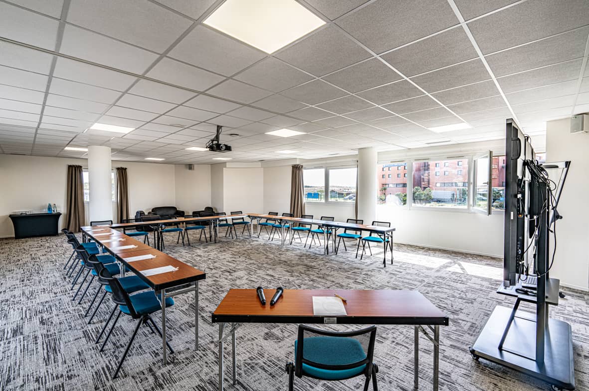 Salle de réunion avec des tables pliantes en bois disposées en U, des chaises en tissu bleu canard à structure métallique noire placées tout autour, un écran numérique mobile sur roulettes au premier plan à droite, un vidéoprojecteur fixé au plafond, un sol en moquette grise à motifs, et de grandes fenêtres avec rideaux beiges laissant voir des bâtiments à l'extérieur sous un ciel lumineux, adaptée à une salle de formation.