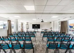 Salle de réunion en configuration amphithéâtre avec des rangées de chaises en tissu bleu canard à structure métallique noire face à un écran numérique noir affichant un logo et un paperboard blanc à l'avant, un sol en moquette grise à motifs, de grandes fenêtres avec rideaux beiges de part et d'autre laissant voir une vue sur la rue à l'extérieur, et une petite table en bois à droite, adaptée à un hôtel pour séminaire.