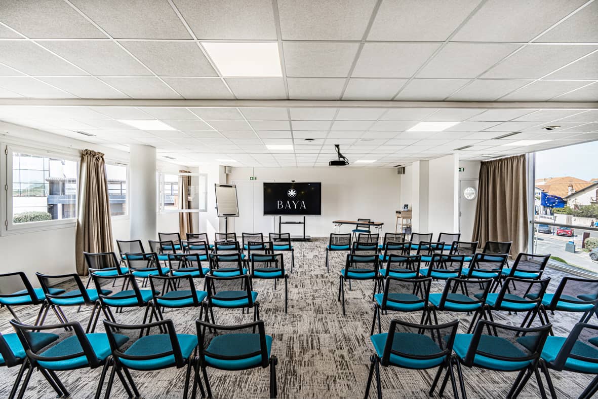 Salle de réunion en configuration amphithéâtre avec des rangées de chaises en tissu bleu canard à structure métallique noire face à un écran numérique noir affichant un logo et un paperboard blanc à l'avant, un sol en moquette grise à motifs, de grandes fenêtres avec rideaux beiges de part et d'autre laissant voir une vue sur la rue à l'extérieur, et une petite table en bois à droite, adaptée à un hôtel pour séminaire.