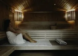 Intérieur de sauna avec des bancs à lattes en bois clair, une femme enveloppée dans une serviette blanche assise sur un tapis rayé gris et blanc à gauche, une serviette rayée pliée sur le banc au centre, et un seau en bois avec une louche posé sur une étagère contre le mur en bois foncé en arrière-plan, éclairé en douceur par des appliques murales, idéal pour un séminaire bien-être.