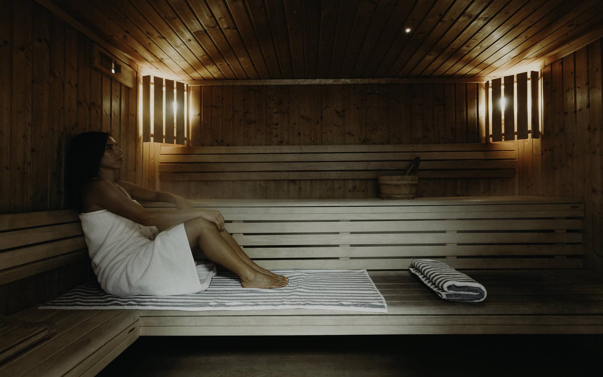 Intérieur de sauna avec des bancs à lattes en bois clair, une femme enveloppée dans une serviette blanche assise sur un tapis rayé gris et blanc à gauche, une serviette rayée pliée sur le banc au centre, et un seau en bois avec une louche posé sur une étagère contre le mur en bois foncé en arrière-plan, éclairé en douceur par des appliques murales, idéal pour un séminaire bien-être.