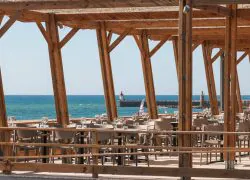 Terrasse de restaurant séminaire en bord de mer avec des tables dressées de verres à vin sous une structure en pergola en bois au premier plan, des chaises en plastique beige alignées au centre, et la mer avec une jetée en pierre et des phares visibles sous un ciel bleu dégagé en arrière-plan.