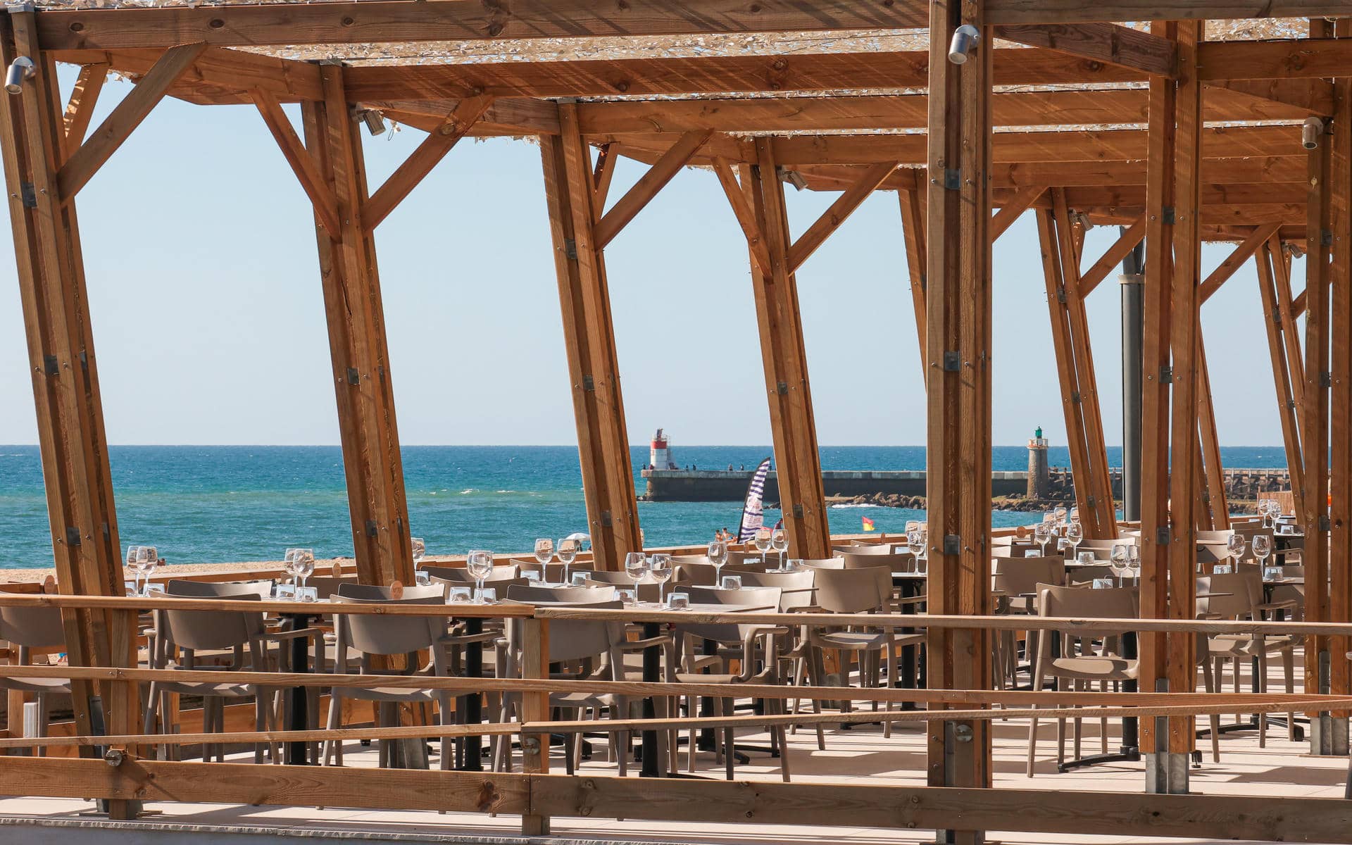 Terrasse de restaurant séminaire en bord de mer avec des tables dressées de verres à vin sous une structure en pergola en bois au premier plan, des chaises en plastique beige alignées au centre, et la mer avec une jetée en pierre et des phares visibles sous un ciel bleu dégagé en arrière-plan.