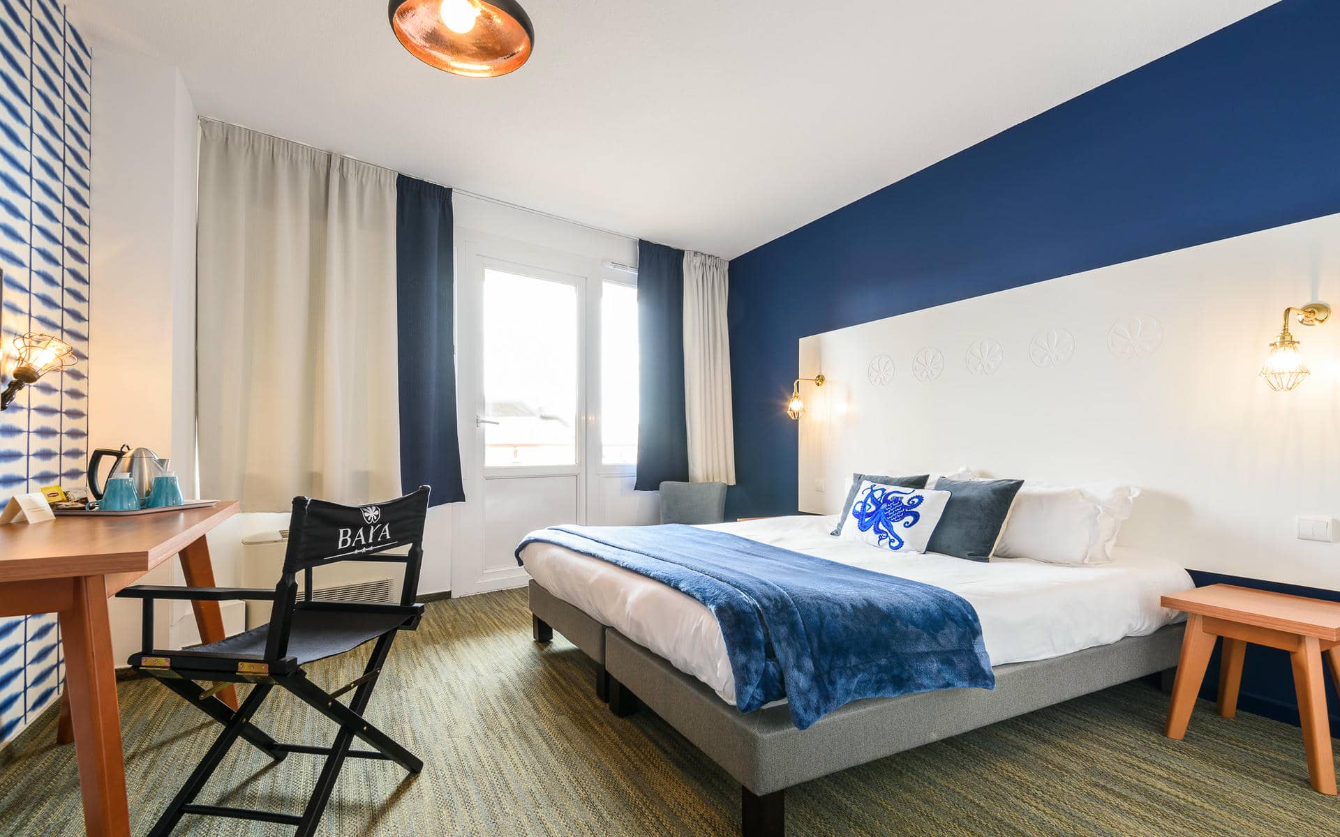 Chambre d'hôtel en bord de mer avec un lit habillé de linge blanc, un coussin motif pieuvre bleu et un jeté bleu marine au centre, une tête de lit blanche gaufrée sous un mur bicolore bleu marine et blanc, un bureau en bois avec bouilloire et chaise de metteur en scène en bois avec logo « BAYA » à gauche, et des fenêtres avec rideaux et porte donnant sur un balcon en arrière-plan.