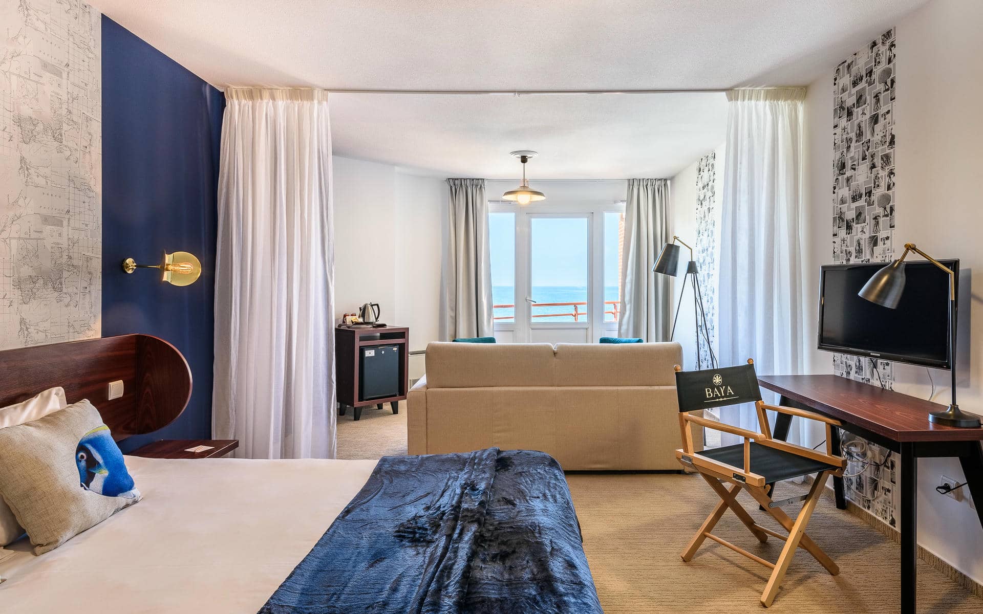 Chambre d'hôtel séminaire en bord de mer avec un lit habillé de linge blanc et un jeté bleu foncé au premier plan, un canapé beige et une chaise de metteur en scène en bois avec logo « BAYA » au centre, un bureau en bois foncé avec lampe métallique à droite près d'une télévision murale, un mur d'accent bleu marine avec papier peint carte ancienne et applique en laiton à gauche, et des portes-fenêtres donnant sur une vue mer en arrière-plan.
