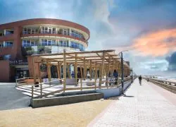 Vue extérieure d'un bâtiment d'hôtel en brique incurvé avec des balcons et une enseigne « Bar Restaurant » à gauche, une pergola en bois surmontée d'un toit en canopée tressée abritant la terrasse où circulent des piétons, une promenade pavée en bord de mer au premier plan avec un cycliste passant à droite, et une plage de sable avec la mer visible au-delà d'un muret sous un ciel spectaculaire au coucher du soleil, adapté à un séminaire en bord de mer.