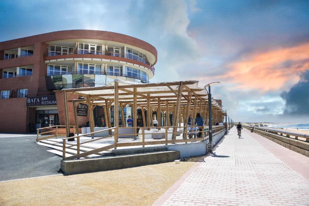 Vue extérieure d'un bâtiment d'hôtel en brique incurvé avec des balcons et une enseigne « Bar Restaurant » à gauche, une pergola en bois surmontée d'un toit en canopée tressée abritant la terrasse où circulent des piétons, une promenade pavée en bord de mer au premier plan avec un cycliste passant à droite, et une plage de sable avec la mer visible au-delà d'un muret sous un ciel spectaculaire au coucher du soleil, adapté à un séminaire en bord de mer.