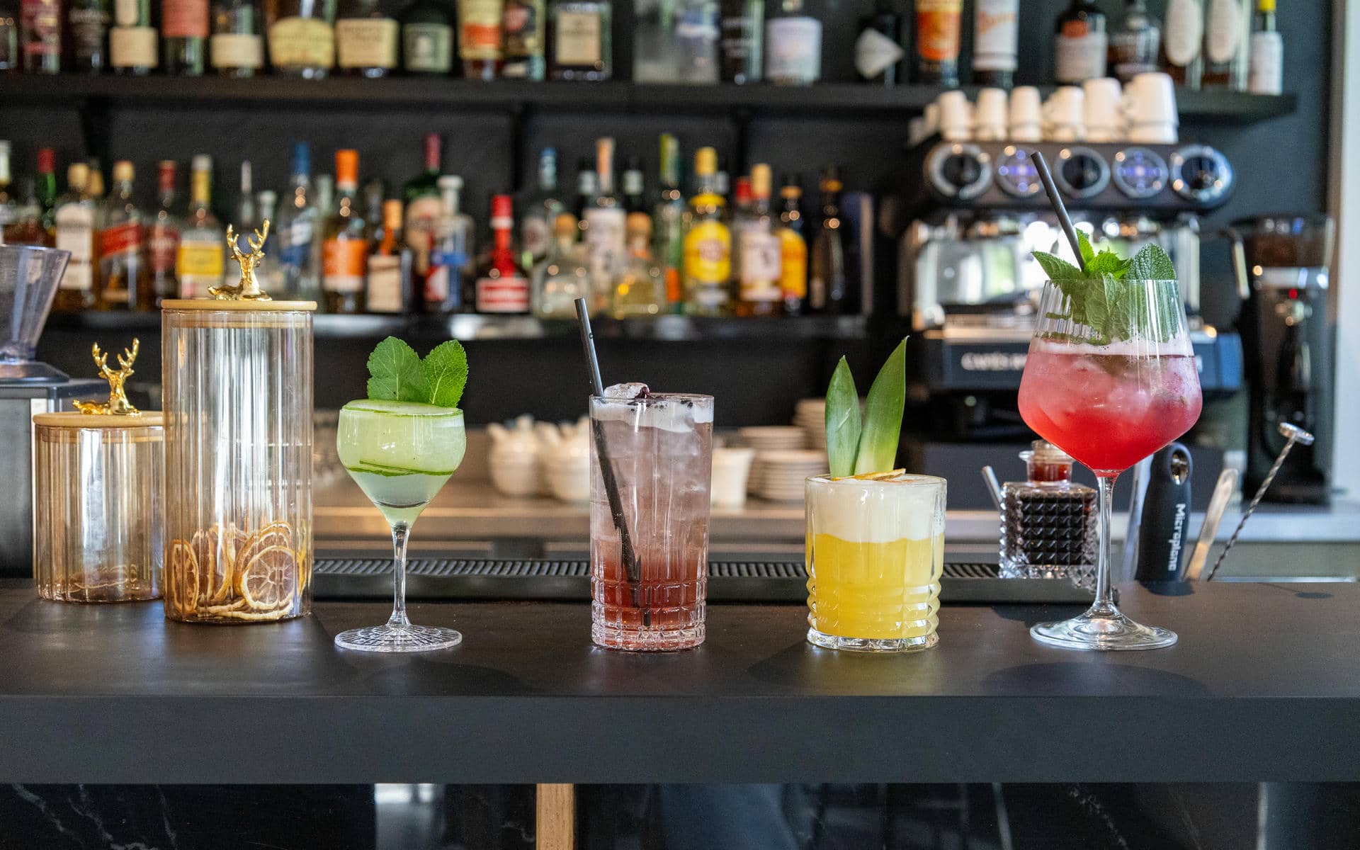 Bar counter set up for an afterwork, featuring five colourful drinks in glasses of different shapes garnished with mint, cucumber and pineapple leaves in the foreground, two glass jars with dried citrus slices to the left, a dark countertop, and shelves lined with bottles and a coffee machine in the blurred background.