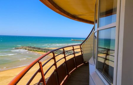 Balcon d'hôtel incurvé avec une balustrade en fer forgé rouge foncé et un plancher en bois à droite, une grande fenêtre reflétant le littoral sur le côté droit, et une vue panoramique sur une plage de sable avec des rochers, une mer turquoise et une jetée au loin sous un ciel bleu dégagé, idéal pour un séminaire en bord de mer.