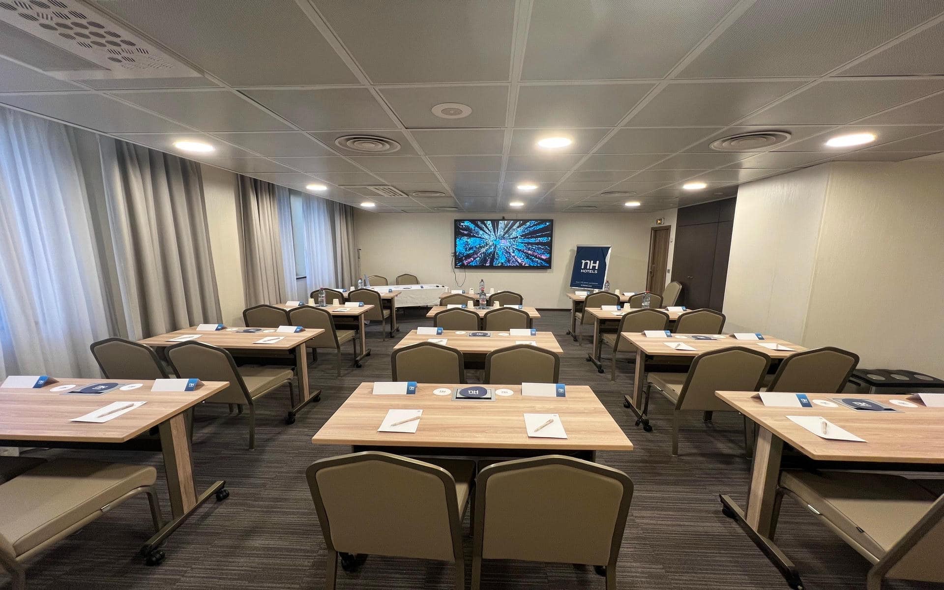Meeting room set classroom-style with wooden tables in rows, each set with a pen, notepad and branded desk sign, beige upholstered chairs on wheels, a wall-mounted screen displaying an aerial forest image and a branded roll-up banner at the front, and beige curtains framing windows on the left, a room laid out for a training room.