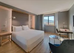 Vue générale d'une chambre d’hôtel avec sol en bois clair pour séminaire en bord de mer, comprenant un lit double à gauche, un fauteuil bleu et une table d'appoint ronde au premier plan à droite, un bureau avec une chaise sous la fenêtre à droite, et des murs gris avec une fenêtre donnant sur un paysage côtier au fond.