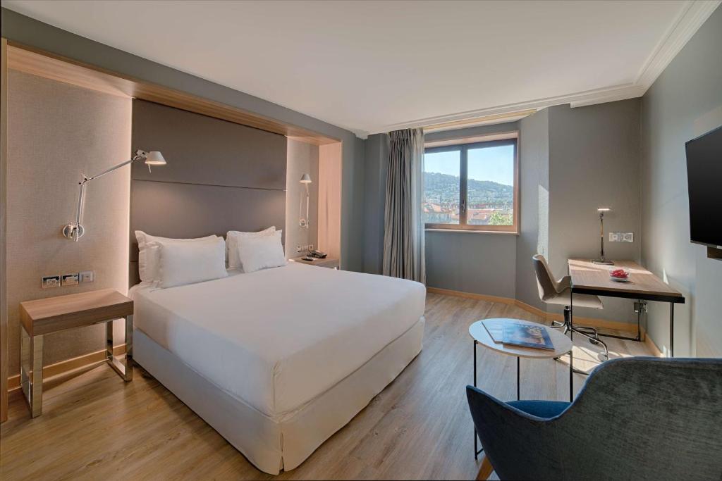 General view of a hotel room with light wood flooring for a seaside seminar, featuring a double bed on the left, a blue armchair and round side table in the foreground on the right, a desk with a chair beneath the window on the right, and grey walls with a window overlooking a coastal landscape at the back.