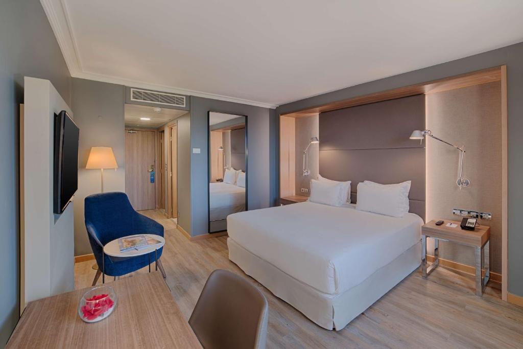 General view of a hotel room with light wood flooring for an urban seminar, featuring a wooden desk with a chair and a glass bowl in the foreground on the left, a blue armchair and round side table on the left, a double bed on the right, a full-length mirror in the centre, and grey walls with bedside tables and reading lamps at the back.
