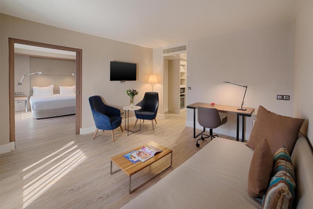 Hotel suite bedroom for seminar hotel with a double bed in the foreground, a wall-mounted reading lamp and light wood headboard on the left, a mirror and wall-mounted television in the centre, and an open doorway leading to a living area with a sofa and dark blue armchair in the background, with light wood flooring.