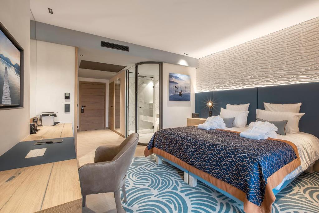 en:: Hotel bedroom for a mountain seminar with a double bed covered by a navy patterned blanket on the right, white pillows against a dark blue headboard, a textured white wall in the background, a light wood desk and chair on the left, and a patterned blue and white carpet on the floor. fr :: Chambre d'hôtel pour un séminaire à la montagne avec un lit double recouvert d'une couverture bleu marine à motifs à droite, des oreillers blancs contre une tête de lit bleu foncé, un mur blanc texturé au fond, un bureau en bois clair et une chaise à gauche, et une moquette bleue et blanche à motifs au sol.
