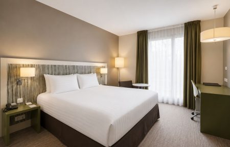 Hotel room for group accommodation with a large white striped bed and dark brown base in the foreground on the left, a striped wood-effect headboard flanked by wall lamps, a green desk with swivel chair on the right, and floor-to-ceiling windows with sheer curtains and olive green drapes revealing trees outside in the background.
