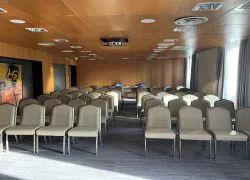 Grande salle de réunion avec rangées de chaises rembourrées beiges et écrues en configuration théâtre séparées par une allée centrale, vidéoprojecteur au plafond face à une table de fond dressée de carafes d'eau, plafond et murs en panneaux de bois, tableau abstrait à gauche, et rideaux gris encadrant de hautes fenêtres à droite, une salle spacieuse configurée pour une location de salle.