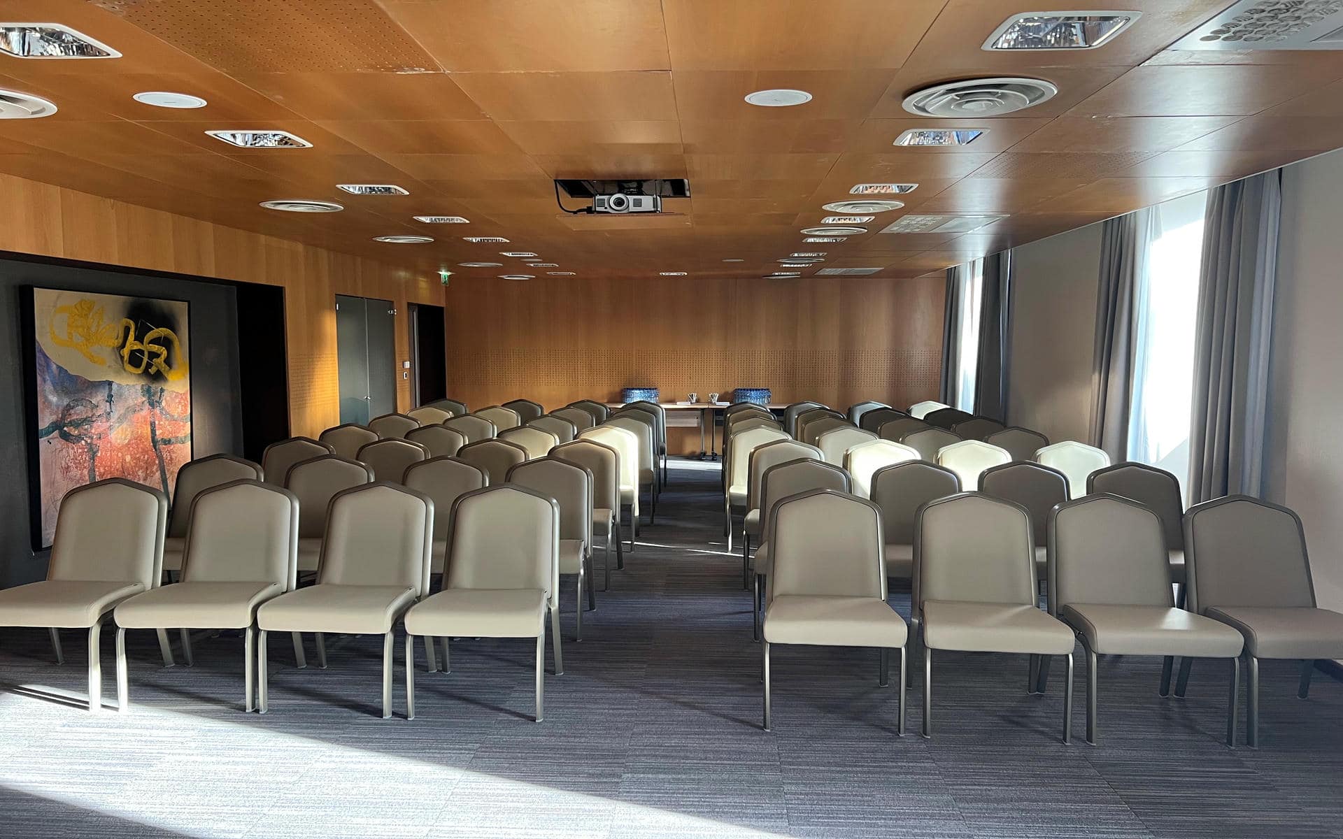 Large meeting room with theatre-style rows of beige and cream upholstered chairs separated by a central aisle, a ceiling-mounted projector above facing a front table with water carafes, a wooden panelled ceiling and walls, an abstract painting on the left, and grey curtains framing tall windows on the right, a spacious room configured for room rental.