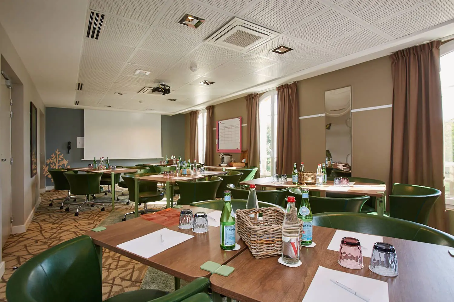 Training room with several wooden tables arranged in rows, green upholstered swivel chairs, water bottles, glasses and wicker baskets in the foreground, a projection screen against a blue-grey wall on the left, tall windows dressed with beige curtains on the right, and patterned brown carpet flooring.