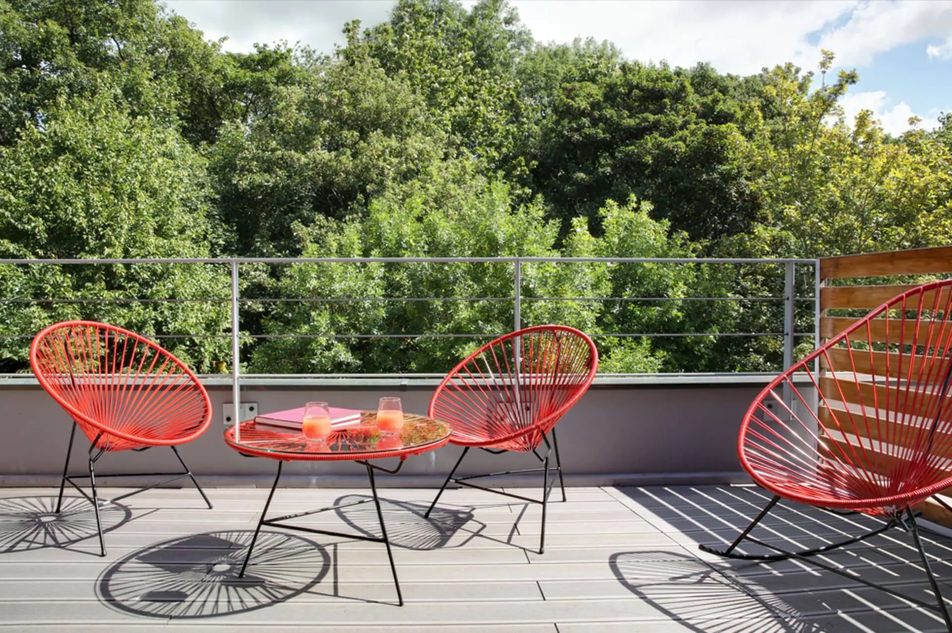 Outdoor terrace ideal for a wellness seminar with a light grey wooden deck in the foreground, red woven acapulco chairs arranged around a round glass table holding drinks and books at the centre, a metal railing along the edge, and dense green trees filling the background under a partly cloudy sky.