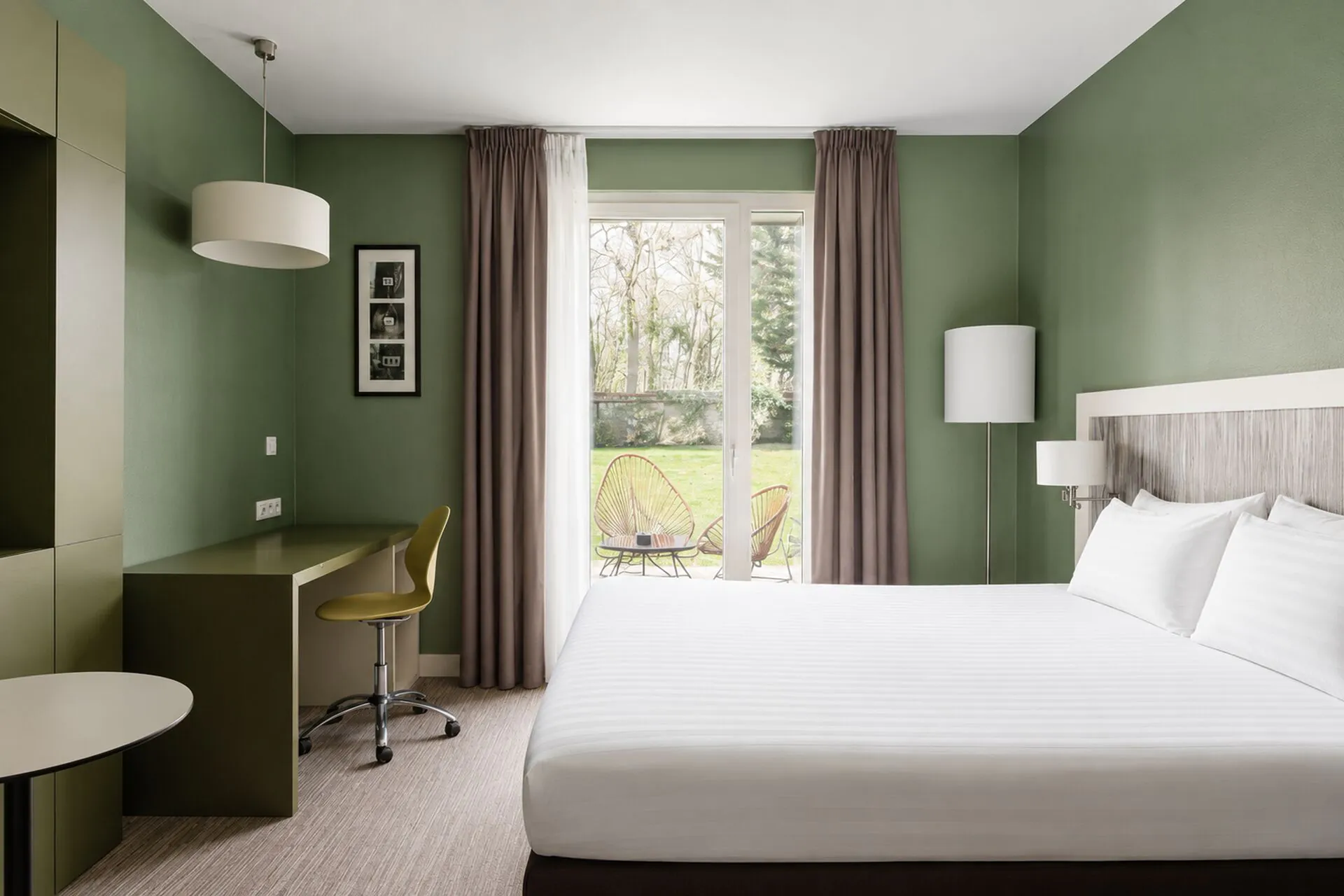 Hotel room for a countryside seminar with a white striped bed on the right, an olive green desk with a mustard yellow swivel chair in the foreground on the left, a framed triptych photo and pendant lamp above, and a floor-to-ceiling window with taupe curtains opening onto a terrace with woven chairs and a lawn in the background.