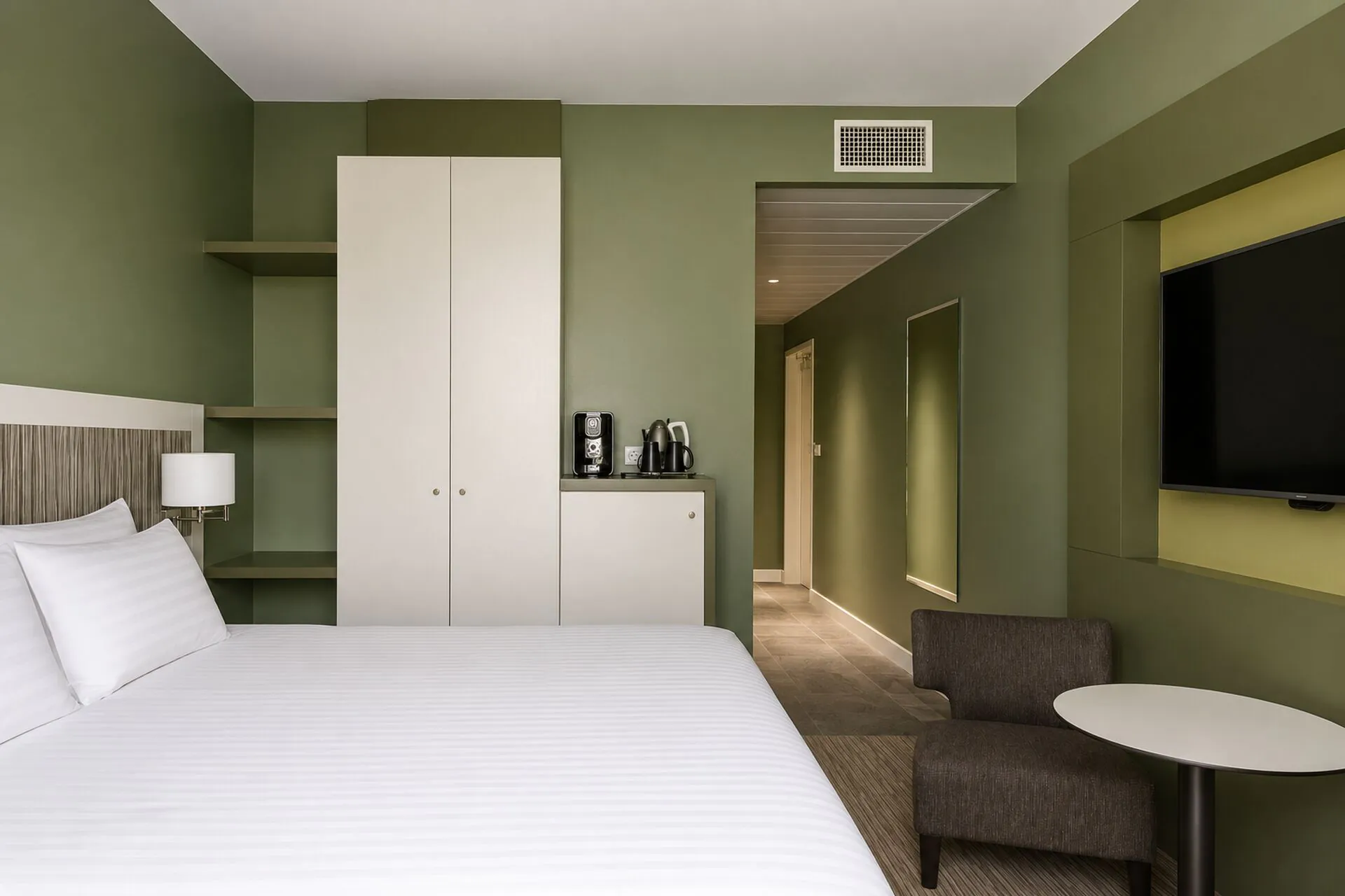 Hotel room for group accommodation with a white striped bed in the foreground on the left, a white wardrobe with wooden shelving and a coffee station on a cabinet against the olive green wall, a wall-mounted flat-screen television on the right, and a dark grey armchair beside a round white side table near the entrance corridor in the background.