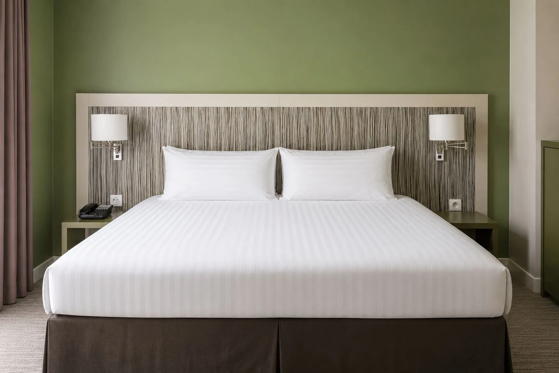 Hotel room for group accommodation with a large white striped double bed and dark brown base in the centre, a striped wood-effect headboard flanked by two white lamp shades on cream bedside tables in the background, a green wall behind the bed, and pink curtains visible on the left.