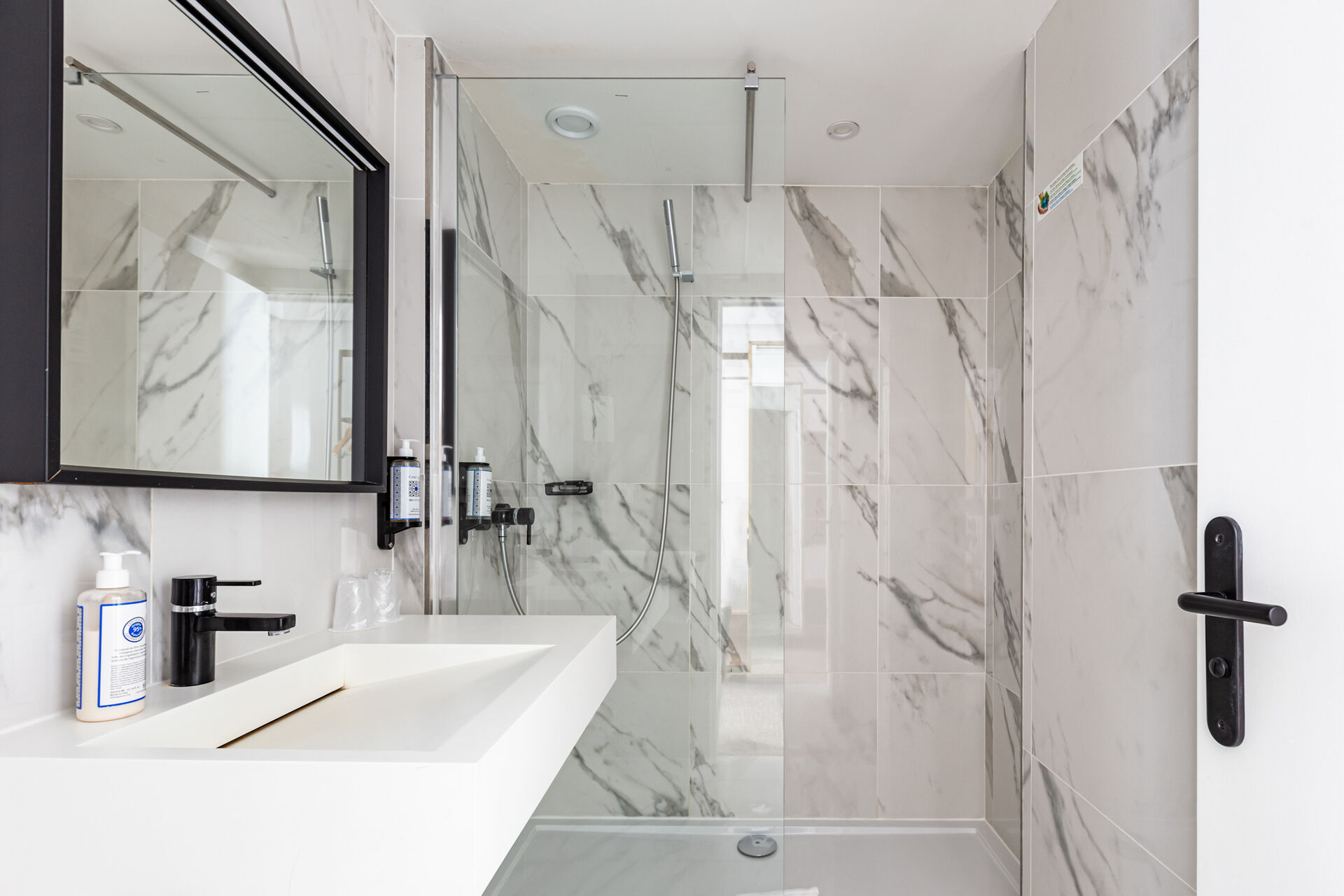 Hotel bathroom for group accommodation with a white marble-effect tiled shower and glass partition in the background, a black-framed mirror and floating white sink with black tap and hand soap bottle on the left, and a black door handle visible on the right against a white door.