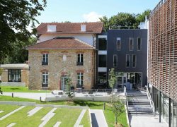 Vue extérieure d'un hôtel pour séminaire en ville, montrant un ancien bâtiment en pierre au toit de tuiles rouges à gauche relié à une extension moderne gris foncé et une façade à claire-voie en bois à droite, une terrasse extérieure avec chaises au centre, et une pelouse paysagée avec dallage géométrique au premier plan sous un ciel bleu.