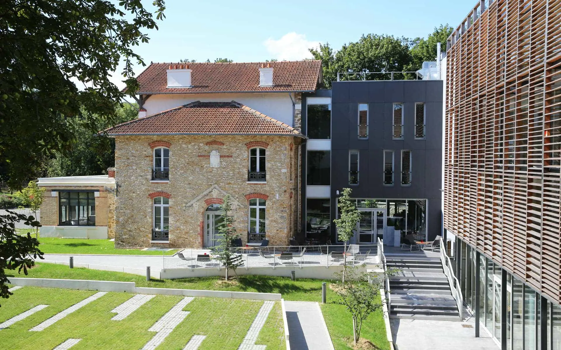 Exterior view of a hôtel pour séminaire at the edge of a city, showing an old stone building with a red-tiled roof on the left connected to a modern dark grey extension and a wooden slatted facade on the right, an outdoor terrace with chairs at the centre, and a landscaped lawn with geometric paving in the foreground under a blue sky.