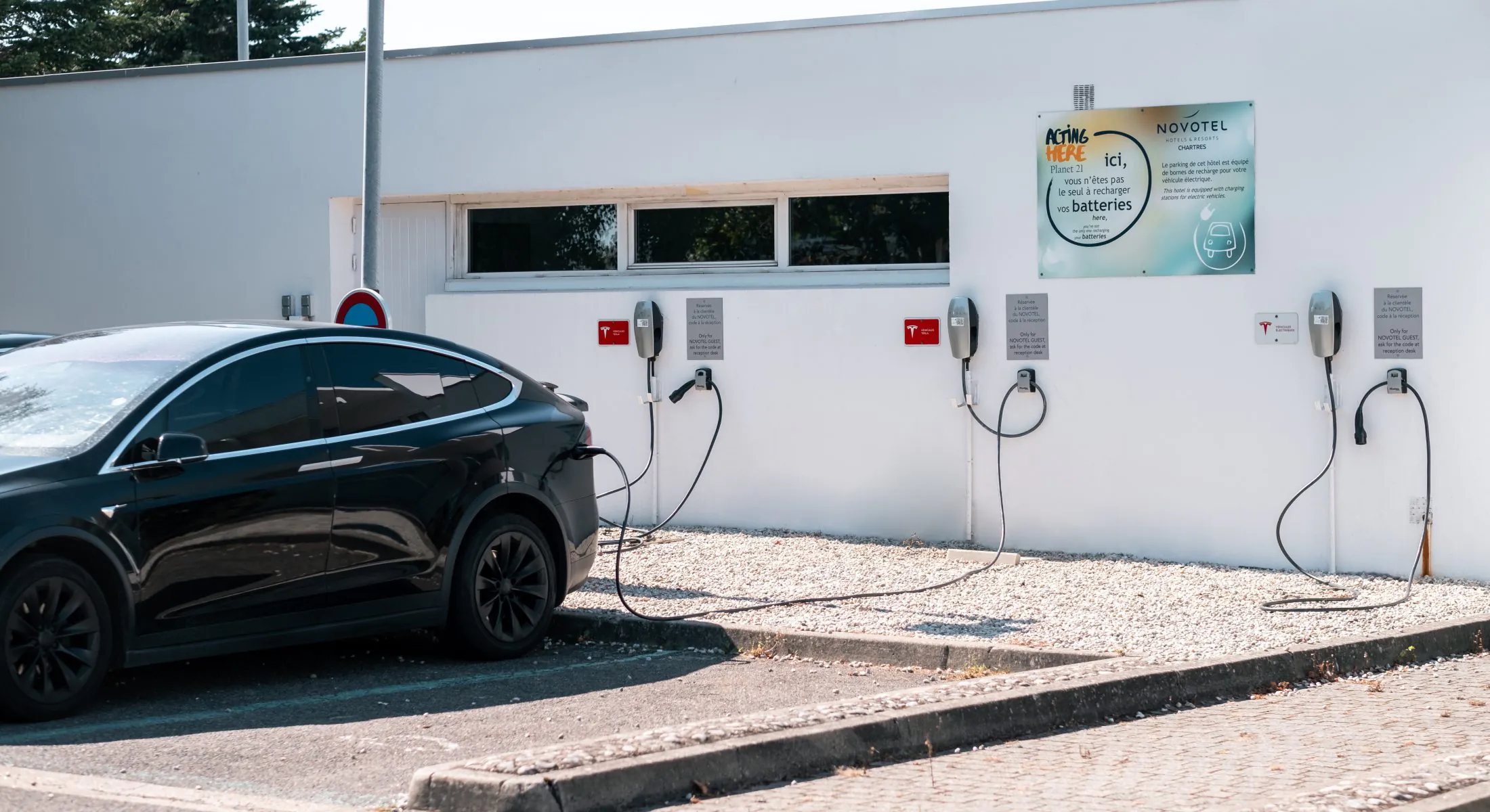 en::Parking area with an electric car charging, featuring Novotel Chartres signage. fr::Parking avec voiture électrique en charge et affichage Novotel Chartres.