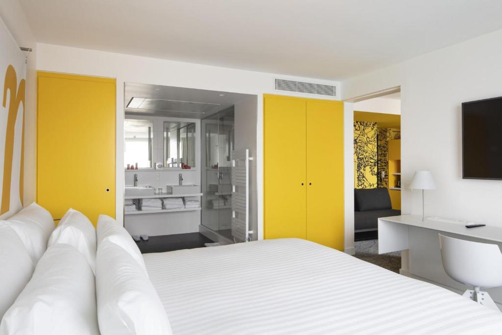 Chambre d'hôtel pour hébergement groupe, avec un grand lit blanc au premier plan, une salle de bain avec douche vitrée et double vasque au centre, des portes de placard jaunes et un bureau blanc avec chaise à droite, et un espace salon avec un mur à motifs graphiques noirs en arrière-plan.