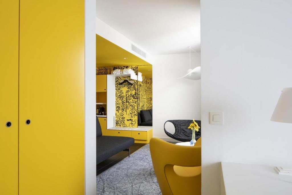 Chambre d'hôtel pour séminaire bien-être, avec un bureau blanc et une lampe à droite au premier plan, un fauteuil jaune et une table ronde au centre, des assises noires, des rangements jaunes et un mur à motifs graphiques noirs en arrière-plan, sur une moquette grise à motifs.