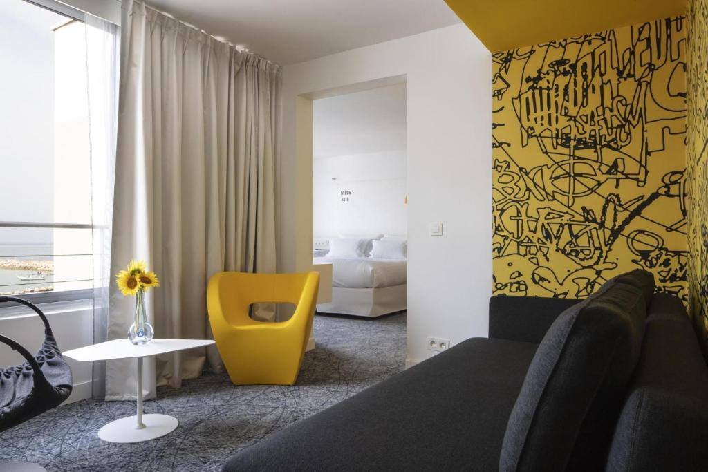 Suite d'hôtel pour hébergement groupe avec un canapé noir au premier plan à droite, un fauteuil jaune et une table basse blanche avec un vase contenant un tournesol à gauche, des rideaux gris toute hauteur le long d'une fenêtre, une ouverture vers la chambre au centre, et un mur décoratif jaune à motifs graphiques noirs sur la droite.