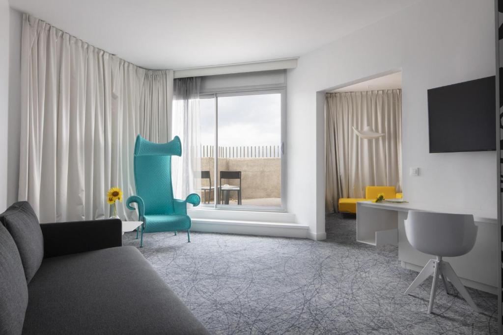 Suite d'hôtel pour hébergement groupe, avec un canapé gris à gauche au premier plan, un fauteuil à haut dossier turquoise face au balcon au centre, un bureau blanc avec chaise et une télévision murale à droite, et une chambre séparée avec une méridienne jaune en arrière-plan sur une moquette grise à motifs.