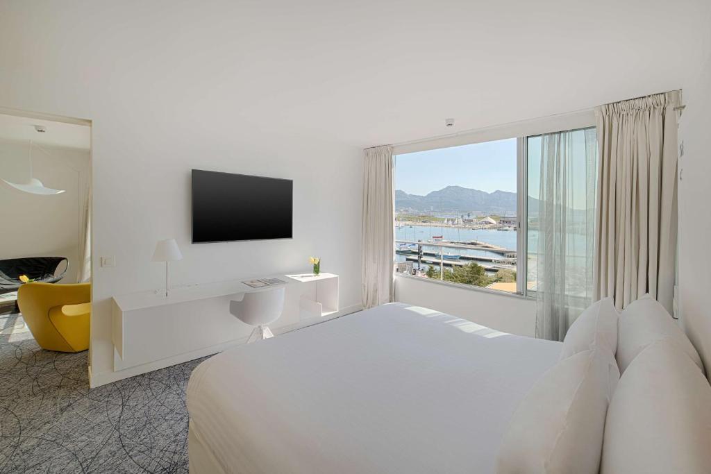Chambre d'hôtel pour séminaire en bord de mer, avec un grand lit blanc au premier plan à droite, un bureau blanc avec chaise et une télévision murale à gauche, une ouverture vers un salon avec un fauteuil jaune en arrière-plan, et une grande fenêtre donnant sur le port de plaisance et les montagnes.