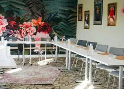 Salle de réunion pour hôtel pour séminaire avec tables blanches disposées en U dressées de bouteilles d'eau, verres et blocs-notes au premier plan, des chaises capitonnées grises sur le pourtour, une grande fresque florale et de fougères recouvrant le mur de gauche, et des affiches illustrées encadrées sur un mur vert pâle à l'arrière-plan.