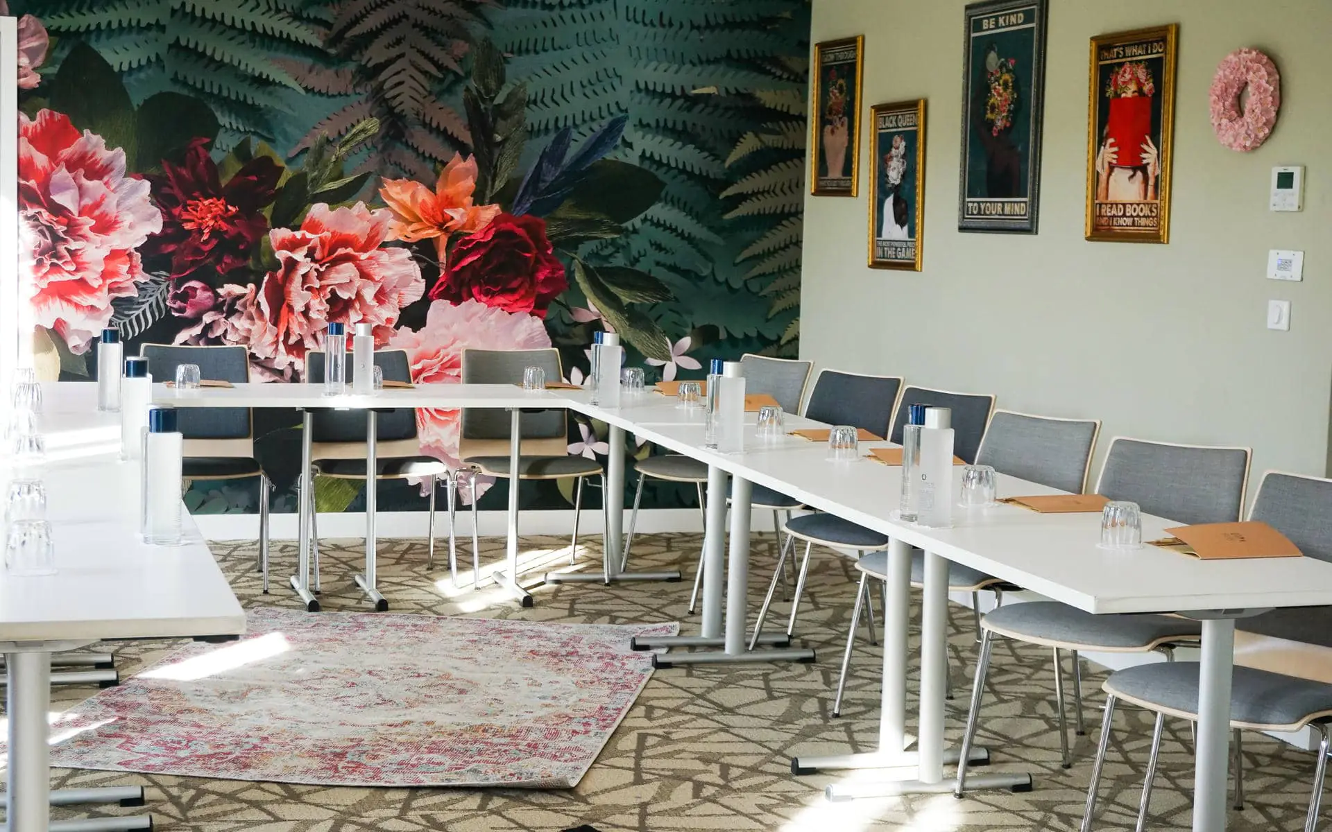 Meeting room for a hôtel pour séminaire with white tables arranged in a U-shape set with water bottles, glasses and notepads in the foreground, grey upholstered chairs on the outer side, a large floral and fern mural covering the left wall, and framed illustrated posters on a pale green wall in the background.